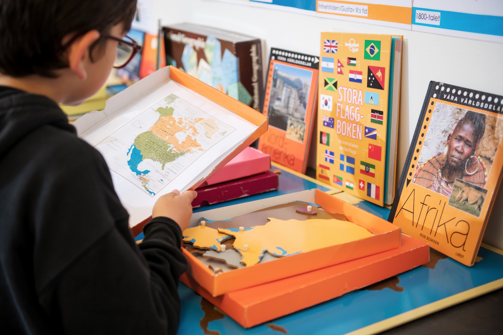 Bild på ett barn som ser på en karta över Nordamerika. På ett bord i bakgrunden finns böckor och pussel om geografi.