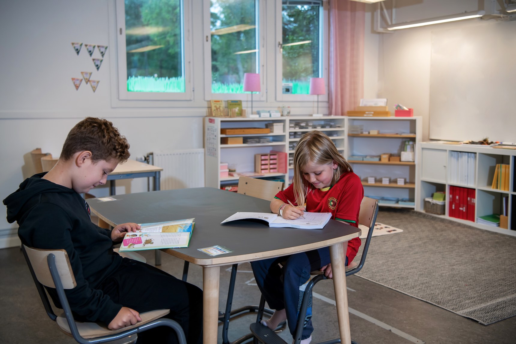 Bild på två barn som sitter vid ett skrivbord. En läser i en bok och en annan antecknar.