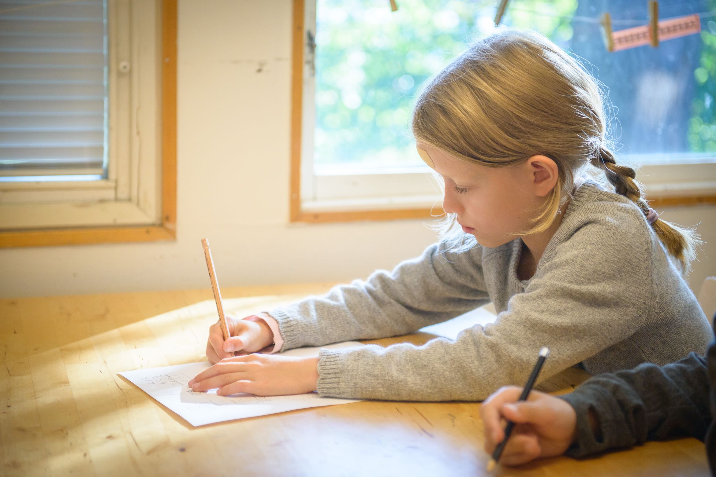 Bild på ett barn som sitter vid ett skrivbord i trä och skriver med blyerts på ett papper medans solen skiner in genom fönstret.